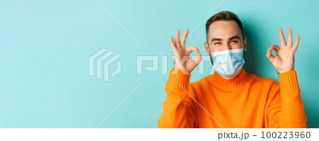 Covid-19, social distancing and quarantine concept. Close-up of satisfied male model in medical mask showing okay signs, praising and agreeing, standing over light blue background Covid-19, social distancing and quarantine concept. Close-up of satisfied male model in medical mask showing okay signs, praising and agreeing, standing over light blue background 100223960