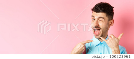 Cheeky and funny guy showing phone gesture, asking to call him and winking, telling dial his number, standing over pink background 100223961