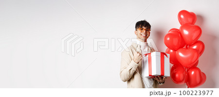 Valentines day and romance concept. Happy and confident boyfriend prepare gift for lover, saying yes and smiling, holding romantic gift, standing near red hearts balloons 100223977