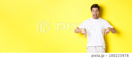 Image of amused handsome guy in white t-shirt, looking and pointing fingers down, standing over yellow background 100223978