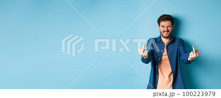Angry caucasian man say fuck you and showing middle fingers, make rude gesture with pissed-off face, standing annoyed on blue background 100223979