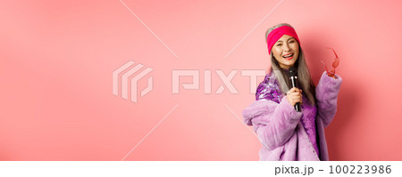 Gorgeous asian senior woman singing karaoke in mic, perform song and looking happy, standing over pink background Gorgeous asian senior woman singing karaoke in mic, perform song and looking happy, standing over pink background 100223986