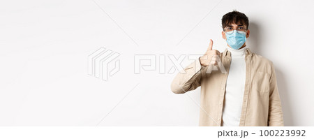 Covid-19, health and real people concept. Satisfied guy in sterile face mask and glasses, showing thumb up in approval, give positive feedback, standing on white background 100223992