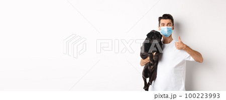 Covid-19, animals and quarantine concept. Young man in medical mask holding cute black pug dog, showing thumb up, like and approve, standing over white background 100223993