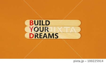 BYD build your dreams symbol. Concept words BYD build your dreams on wooden stick on a beautiful orange table orange background. Business and BYD build your dreams concept. Copy space. 100225914
