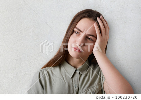 A portrait of thoughtful girl with long dark hair and big brown eyes wearing grey T-shirt having a sad expression holding her hand on her head. A tired girl having deep thoughts about something 100230720