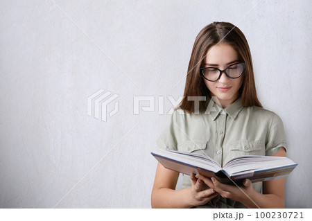 Education and studying. A portrait of a clever beautiful girl with long dark hair wearing glasses holding and reading a hardcover book on white background. An attentive reading of the book 100230721