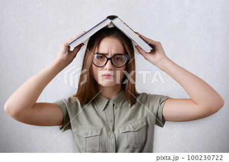A closeup portrait of pretty young schoolgirl wearing glasses on white background holding her book over her head being tired of reading and having fun. Girl making wry face with a book overhead 100230722