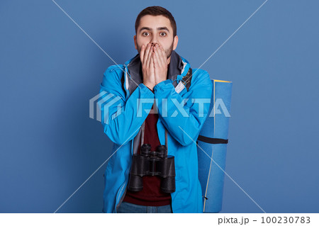 Image of impressed confused handsome explorer covering his mouth with both hands, looking directly at camera, wearing blue jacket, dark blue sweatshirt, having all equipment for hiking alone. 100230783