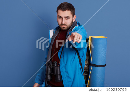 Close up portait of young caucasian man dresses maroon shirt and blue jacket, pointing at camera with his fore finger, bearded guy with serious facial expression, posing isolated over studio wall. 100230796