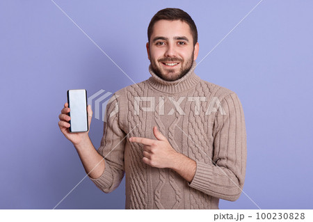 Portrait of happy man dresses in beige knitted sweater, standing isolated over lilac background, holding mobile phone with blank screen, pointing at device. Copy space for advertisment or promotion. 100230828