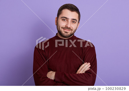 Portrait of positive magnetic adorable man standing isolated over lilac background in studio, looking directly at camera, having folded arms, feeling comfortable, spending leisure time alone. 100230829