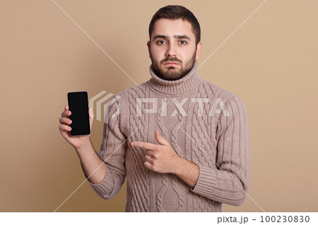 Close up portrait of nice calm handsome young man holding smartphone in one hand, pointing at it with forefinger, being upset, standing isolated over beige background in studio. Choice concept. Close up portrait of nice calm handsome young man holding smartphone in one hand, pointing at it with forefinger, being upset, standing isolated over beige background in studio. Choice concept. 100230830