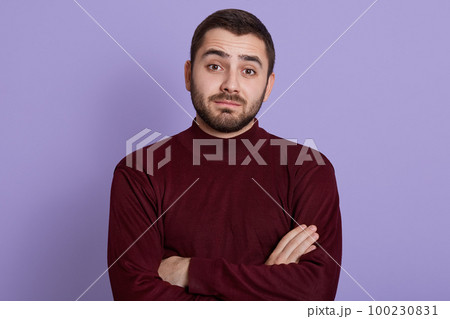 Thoughtful young man with skeptic, doubtful, distrustful look posing against lilac background with folded hands, wearing burgundy sweater, bearded puzzled male. 100230831