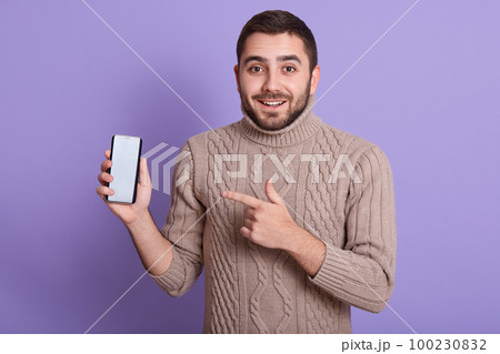 Horizontal shot of young man pointing at blanck screen of his phone, looks happy, looking smiling at camera, bearded man wearing beige sweater, male posing isolated over lilac studio background. Horizontal shot of young man pointing at blanck screen of his phone, looks happy, looking smiling at camera, bearded man wearing beige sweater, male posing isolated over lilac studio background. 100230832
