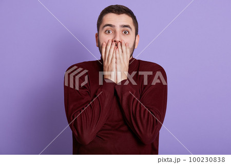 Portrait of scared impressed man with beard standing isolated over lilac background in studio, covering half of his face with hands, opening his eyes widely, being shocked. Emotions concept. 100230838