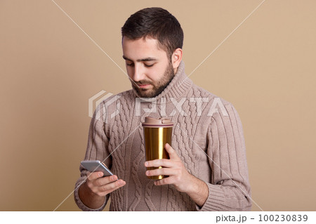 Image of concentrated busy attractive man holding his smartphone and thermal mug in one hand, using his cellphone, drinking coffee, having lunch, communicating with others. Communication concept. 100230839