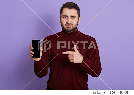 Horizontal shot of young sad man with disappointed facial expression, making gesture, holding smartphone, pointing at it with forefinger, being unsatisfied with choice and purchase. Tech concept. Horizontal shot of young sad man with disappointed facial expression, making gesture, holding smartphone, pointing at it with forefinger, being unsatisfied with choice and purchase. Tech concept. 100230890