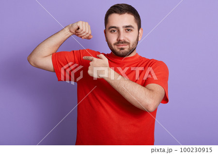 Close up portrait of happy strong man dresses casual red t shirt showing his biceps on arm and pointing with index finger, male looking at camera while standing isolated over purple background. Close up portrait of happy strong man dresses casual red t shirt showing his biceps on arm and pointing with index finger, male looking at camera while standing isolated over purple background. 100230915