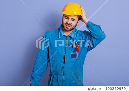 Studio shot of handsome young bearded Caucasian mechanic wearing blue work clothes and yellow hard hat, stands touching his head, having painful look, feeling exhausted after hard working day. Studio shot of handsome young bearded Caucasian mechanic wearing blue work clothes and yellow hard hat, stands touching his head, having painful look, feeling exhausted after hard working day. 100230970