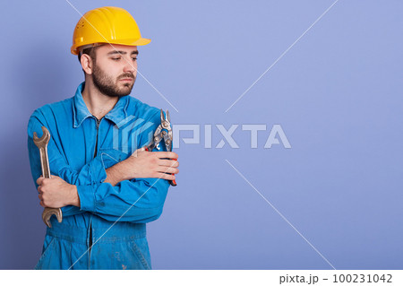 Close up portrait of young man wearing blue working clothes and yellow protective helmet, posing with wrench tools in hands isolated over blue studio background. Copy space for advertisment. 100231042