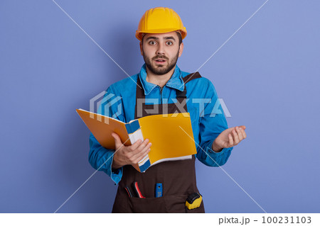 Astonished shocked young builder in yellow helmet standing with mouth opened and being very surprised, holding yellow paper folder in hands, sees mistake in drawing. Astonished shocked young builder in yellow helmet standing with mouth opened and being very surprised, holding yellow paper folder in hands, sees mistake in drawing. 100231103