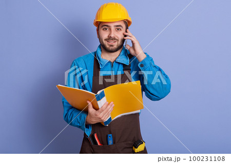 Close up portrait of young attractive bearded builder having conversation via phone and holding paper folder in hands, dresses blue shirt, brown apron, yellow helmet. Buildingand engineering concept. 100231108