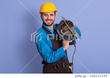 Horizontal photo of good looking handsome happy worker holding tool in both hands, looking directly at camera, smiling sincerely, wearing brown apron, blue overall and yellow helmet. Work concept. Horizontal photo of good looking handsome happy worker holding tool in both hands, looking directly at camera, smiling sincerely, wearing brown apron, blue overall and yellow helmet. Work concept. 100231145