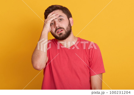 Studio shot of tensed bearded young man touches his forehead and having headache, wearing red casual t shirt, standing and looking at camera isolated over yellow background. Healthy care concept. 100231204