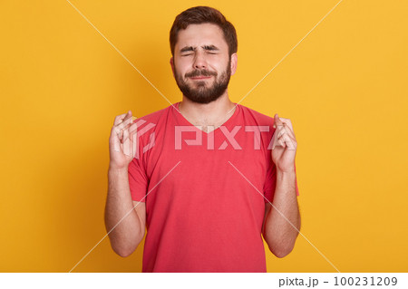 Close up portrait of handsome young man wearing red t shirt keeping eyes closed and fingers crossed while standing against yellow background, waiting results of exam, wants his dream comes true. Close up portrait of handsome young man wearing red t shirt keeping eyes closed and fingers crossed while standing against yellow background, waiting results of exam, wants his dream comes true. 100231209