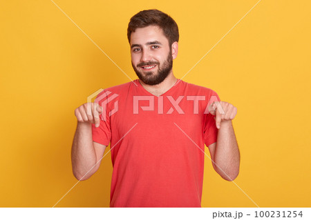 Horizontal shot of young beraded male, looking directly at camera, wearing casual bright red t shirt, standsing against white studio wall, pointing fore fingers down. Copy space for advertisment. 100231254