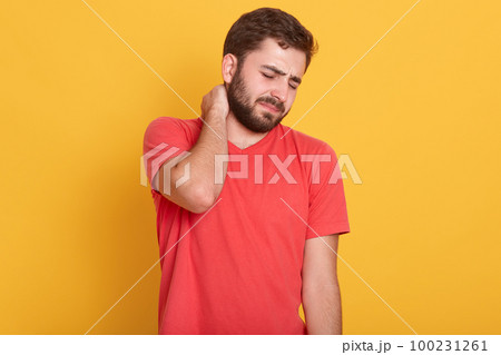 Image of attractive bearded male feeling pain in neck, frustrated young man wearing red casual t shirt, touching his neck and expressing negativity while standing against yellow studio background. 100231261