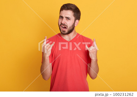 Close up portrait of man showing rock gesture against yellow background, wearing red casual t shirt, screaming somthing loudly, bearded guy, looking directly at camera. People emotions concept. 100231262