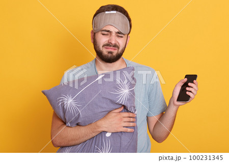 Horizontal indoor shot of sleepy miserable man wearing sleeping mask and t shirt, holding pilow and smartphone, waking up, putting off alarm clock, standing isolated over yellow background in studio. Horizontal indoor shot of sleepy miserable man wearing sleeping mask and t shirt, holding pilow and smartphone, waking up, putting off alarm clock, standing isolated over yellow background in studio. 100231345