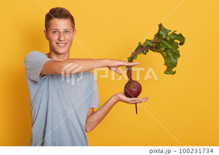 Indoor studio shot of handsome model standing isolated over yellow background, holding raw vegetable in one hand, showing size, making gesture, smiling sincerely. People and healthy food concept. Indoor studio shot of handsome model standing isolated over yellow background, holding raw vegetable in one hand, showing size, making gesture, smiling sincerely. People and healthy food concept. 100231734