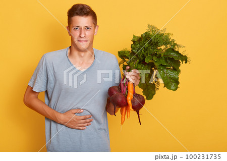 Horizontal shot of handsome male holding carrotsa and beets in hand, posing in casual grayt shirt with fresh vegetables, keeps hand on stomach, feels hunger. Raw food diet and healthy eating concept. Horizontal shot of handsome male holding carrotsa and beets in hand, posing in casual grayt shirt with fresh vegetables, keeps hand on stomach, feels hunger. Raw food diet and healthy eating concept. 100231735