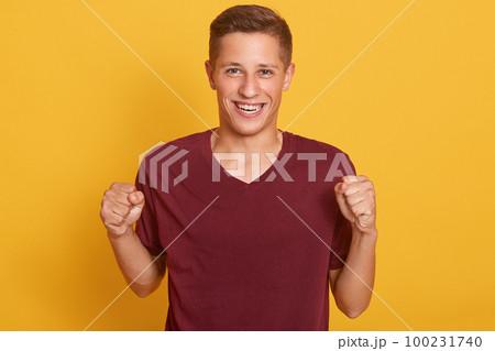 Happy positive excited young man clenching fists and screaming, attractive guy wearing maroon casual t shirt glad to hear good news, celebrating his victory or success, isolated over yellow background 100231740