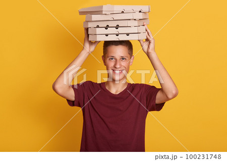 Close up portrait of young cheerful delivery man with red t-shirt standing,holding stack of cardboard pizza boxes on grey background. Indoor, studio shot, isolated, copy space. Close up portrait of young cheerful delivery man with red t-shirt standing,holding stack of cardboard pizza boxes on grey background. Indoor, studio shot, isolated, copy space. 100231748