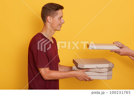Profile of cheerful pleasant young boy holding cardboard boxes with pizza, unknown hands putting one more box, smiling sincere, standing isolated over yellow background in studio. Delivery concept Profile of cheerful pleasant young boy holding cardboard boxes with pizza, unknown hands putting one more box, smiling sincere, standing isolated over yellow background in studio. Delivery concept 100231765