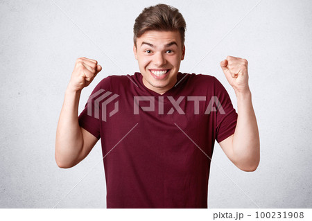 Happy joyful strong male clenches fists and teeth, dressed in casual t shirt, celebrates his victory and success, isolated over white background. Glad cheerful man youngster gestures indoor. 100231908