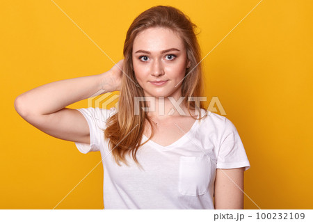 Closeup portrait of positive energetic young lady touching her hair with one hand, looking directly at camera, having pleasant facial expression, standing isolated over yellow background, being calm. 100232109