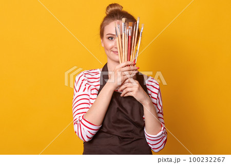 Horizontal shot of girl artist holds brushes in hands and hides behind it, lady dresses stripes casual shirt and brown apron, blonde woman with bunch, posing isolated over yellow studio background. 100232267
