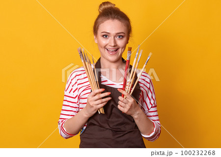 Portrait of gorgeous female artist working on art projects, attractive woman wearing striped casual shirt and brown apron, charming blonde girl has happy facial expression, holding brushes in hands. 100232268