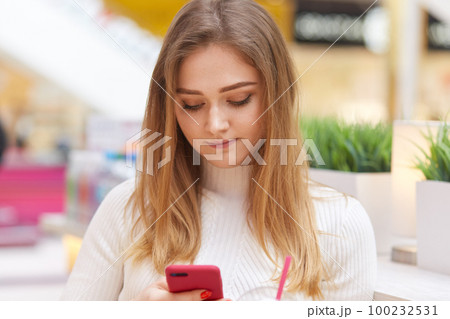Teenage girl with long hair, focused in screen of cell phone, reads news in internet, surfes social networks, reads post under photo, poses against blurred background indoor, has spare time. Teenage girl with long hair, focused in screen of cell phone, reads news in internet, surfes social networks, reads post under photo, poses against blurred background indoor, has spare time. 100232531