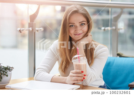 Image of cute adorable femal looking directly at camera, having lunch in cafeteria, holding plastic cup of milkshake, putting laptop on table, looks delighted and positive, wearing casual sweater. 100232601
