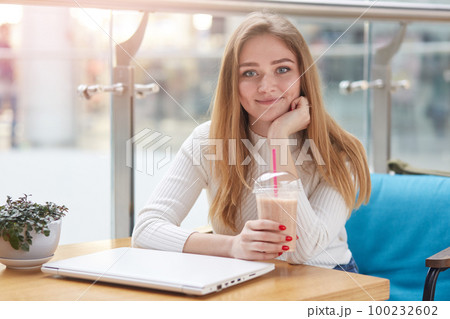 Close up portrait of beautiful young caucasian woman with long blonde hair sitting in cafe, drinking milk cocktail, having rest after online working on laptop, looking smiling directly at camera. 100232602