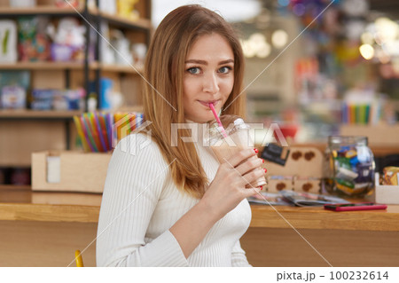 Photo of attractive female drinks milk cocktail with straw, poses in bar, wears white jumper, looks directly at camera, enjoys its taste, has spare time after work or classes. People and lifestyle 100232614