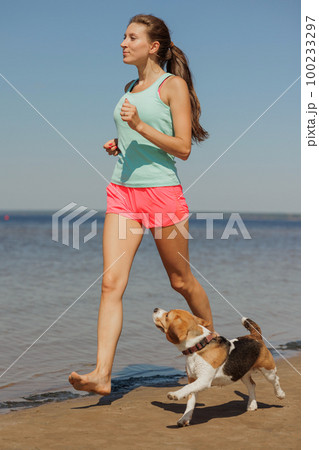 athletic woman and beagle dog run along sandy beach on seashore in morning. outdoor 100233297
