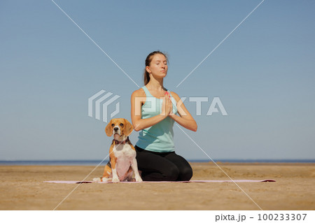 woman and dog meditate on yoga mat on ocean in morning. sports outdoor for mental health 100233307