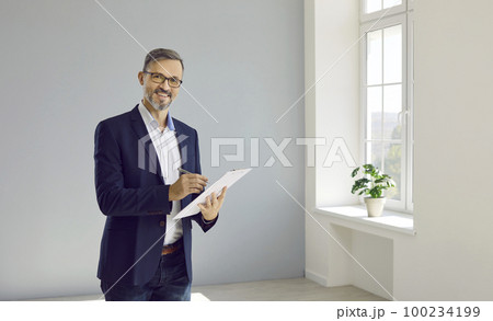 Smiling realtor man fills out documents in clipboard while standing in apartment for sale. 100234199